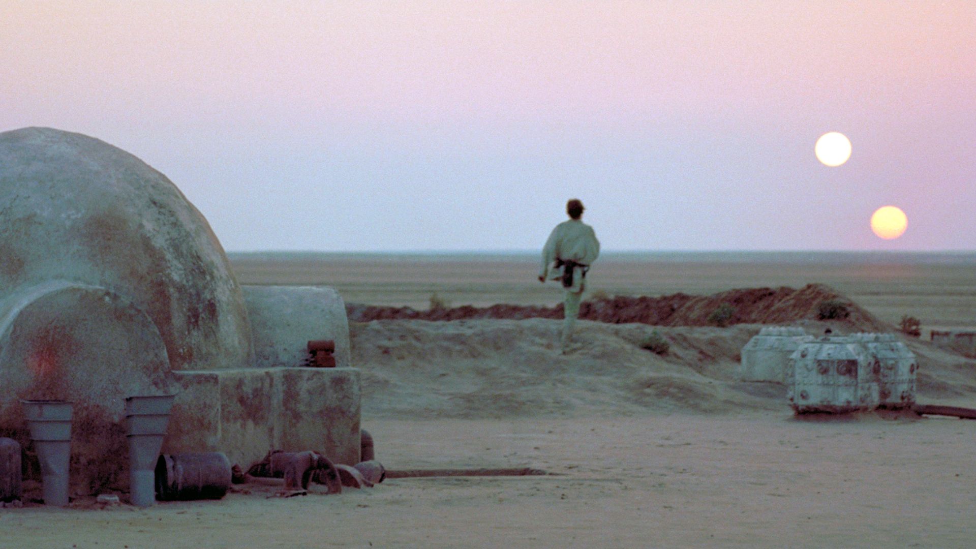 A man with his back to the viewer watches Tatooine's binary sunset at his homestead